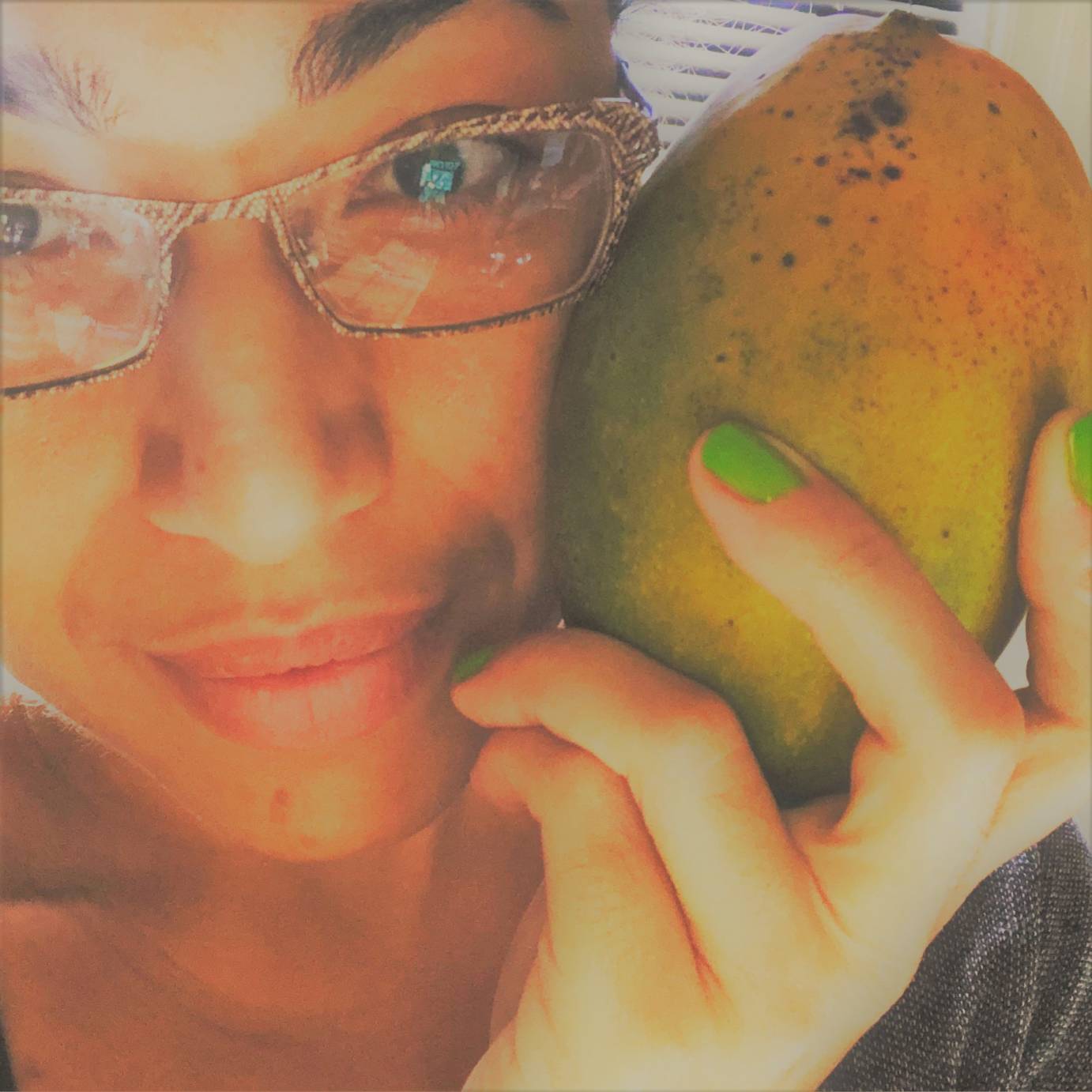 A close up of a Black woman's facing, wearing animal-print-like glasses holding with bright green lacquered fingers an overripe mango against her left cheek